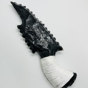May include: A black obsidian knife with a jagged blade and a handle wrapped in white cord. The blade has a textured surface and a sharp point. The knife is displayed against a white background.