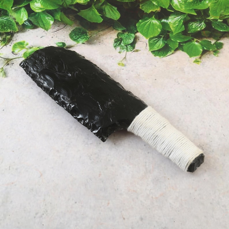 May include: A black obsidian knife with a white wrapped handle. The blade is a dark, glossy black with a rough, textured edge. The handle is tightly wrapped with white cord, providing a secure grip. The knife is displayed on a light-colored surface.