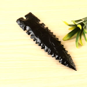 May include: A black obsidian arrowhead with a serrated edge. The arrowhead is on a light wood surface.