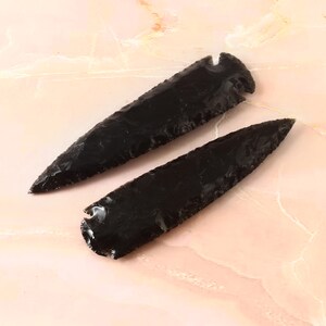 May include: Two black obsidian arrowheads, one larger than the other, are displayed on a pink surface. The arrowheads are sharp and have a pointed tip.