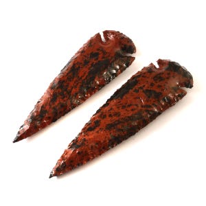 Puede incluir: Dos puntas de flecha rojas y negras hechas de sílex. Las puntas de flecha son puntiagudas y tienen una textura rugosa.