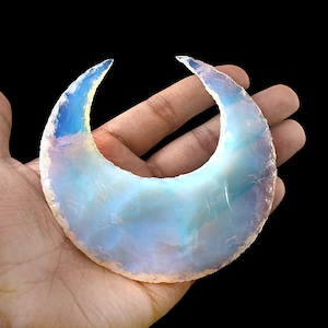 May include: A crescent-shaped, opalescent gemstone with a polished surface. The stone displays a mix of pale blue and white hues, with a translucent quality. The edges are slightly rough, and the overall shape resembles a stylized moon.