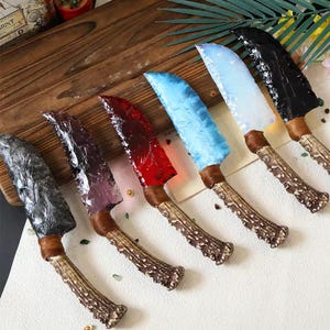 Könnte beinhalten: Eine Sammlung von sieben dekorativen Messern mit Klingen in verschiedenen Farben, darunter Schwarz, Grau, Rot, Blau und Weiß. Jedes Messer hat einen braunen Griff mit einem strukturierten, geweihartigen Aussehen. Die Klingen haben ein einzigartiges, strukturiertes Aussehen.