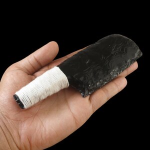 May include: A black obsidian knife with a white wrapped handle. The obsidian blade has a rough, textured surface. The handle is wrapped with white string. The knife is held in an open hand against a black background.