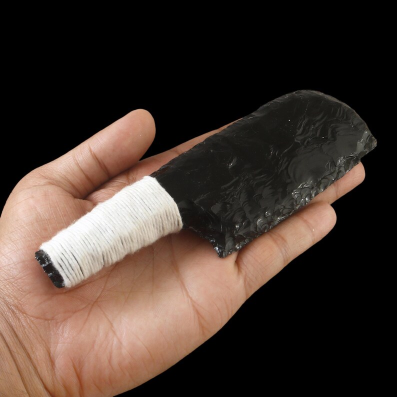May include: A black obsidian knife with a white wrapped handle. The obsidian blade has a rough, textured surface. The handle is wrapped with white string. The knife is held in an open hand against a black background.