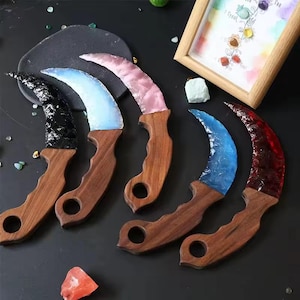 May include: A collection of karambit knives with wooden handles and curved blades in various colors, including black, blue, pink, and red. The blades have a textured, crystalline appearance. The knives are displayed on a dark surface.