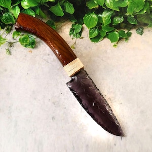 Puede incluir: Un cuchillo artesanal con un mango de madera oscura y una hoja teñida de púrpura. El mango es curvo y está envuelto con un cordón de color natural cerca de la hoja. La hoja tiene un borde afilado y una superficie texturizada.