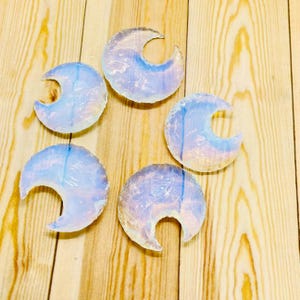 May include: Five opalescent crescent moon-shaped pendants. Each pendant has a translucent, iridescent quality with shades of blue and white. The pendants are arranged on a light brown wooden surface.
