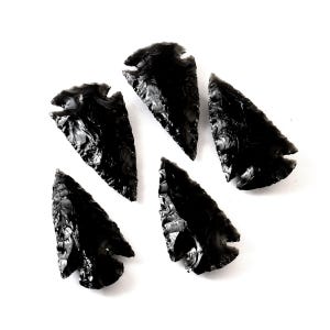 Puede incluir: Cinco puntas de flecha de obsidiana negra dispuestas sobre un fondo blanco. Cada punta de flecha tiene forma triangular con base dentada y punta afilada. La obsidiana presenta una superficie brillante y reflectante.