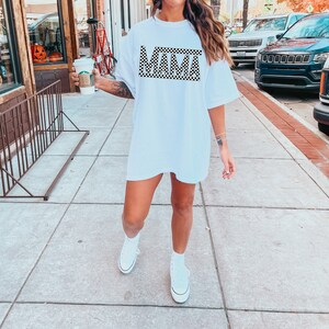 Puede incluir: Camiseta blanca de gran tama&ntilde;o con la palabra "MAMA" en un patr&oacute;n de cuadros blanco y negro. La camiseta se usa con zapatillas altas blancas. La imagen est&aacute; tomada al aire libre en una acera.