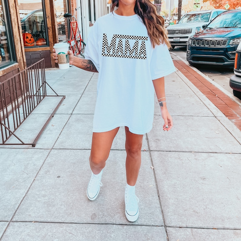 Puede incluir: Camiseta blanca de gran tama&ntilde;o con la palabra "MAMA" en un patr&oacute;n de cuadros blanco y negro. La camiseta se usa con zapatillas altas blancas. La imagen est&aacute; tomada al aire libre en una acera.