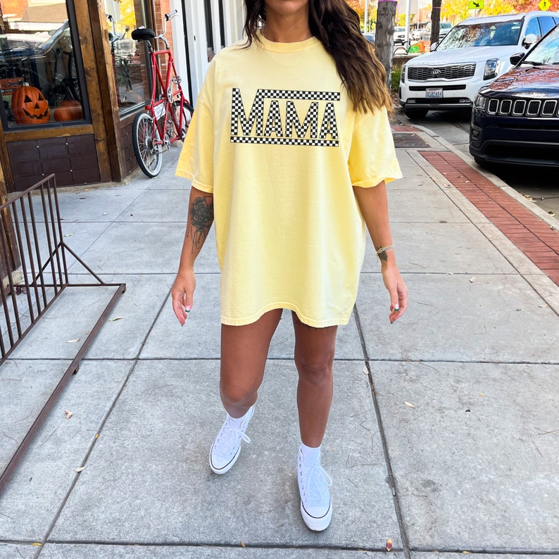 Puede incluir: Camiseta amarilla clara con la palabra "MAMA" en letras negras, enmarcada por un patr&oacute;n de cuadros blancos y negros. La camiseta se lleva con zapatillas blancas.