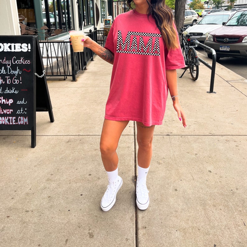 Puede incluir: Camiseta roja con la palabra "MAMA" en un dise&ntilde;o a cuadros blanco y negro. La camiseta se usa con zapatillas y calcetines blancos. La imagen se toma al aire libre en un d&iacute;a soleado.