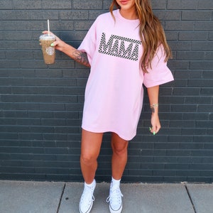 Puede incluir: Una camiseta rosa claro de gran tama&ntilde;o con la palabra "MAMA" en un dise&ntilde;o a cuadros blanco y negro. La camiseta se usa con zapatillas blancas. La persona sostiene un vaso de pl&aacute;stico transparente con una pajita.