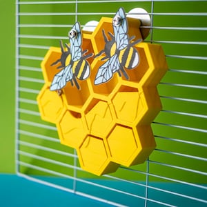 Pode incluir: Brinquedo amarelo em forma de favo de mel com dois recortes de abelhas anexados. O brinquedo é feito de plástico e tem uma superfície texturizada. É projetado para ser pendurado em uma gaiola.