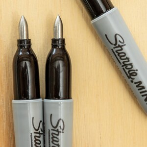 K&ouml;nnte beinhalten: Nahaufnahme von drei Sharpie Mini Markern. Zwei Marker haben ihre silbernen Spitzen freigelegt, mit schwarzen Tintenbeh&auml;ltern und grauen Geh&auml;usen. Der dritte Marker ist teilweise sichtbar und zeigt das graue Geh&auml;use mit dem Sharpie Mini-Logo. Der Hintergrund ist eine helle Holzoberfl&auml;che.