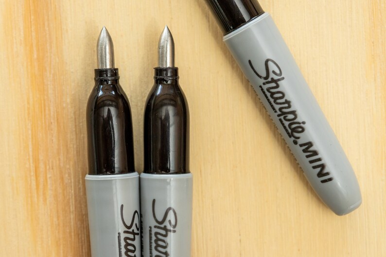 K&ouml;nnte beinhalten: Nahaufnahme von drei Sharpie Mini Markern. Zwei Marker haben ihre silbernen Spitzen freigelegt, mit schwarzen Tintenbeh&auml;ltern und grauen Geh&auml;usen. Der dritte Marker ist teilweise sichtbar und zeigt das graue Geh&auml;use mit dem Sharpie Mini-Logo. Der Hintergrund ist eine helle Holzoberfl&auml;che.