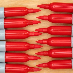 May include: A close-up of ten red and grey Sharpie permanent markers lined up in a row. The markers are all facing the same direction with the tips pointing towards the top of the image. The markers are all the same size and shape.
