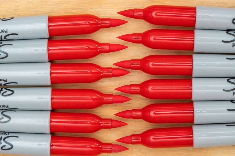 May include: A close-up of ten red and grey Sharpie permanent markers lined up in a row. The markers are all facing the same direction with the tips pointing towards the top of the image. The markers are all the same size and shape.