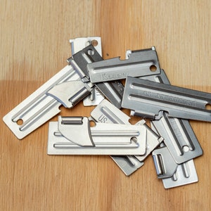 May include: A close-up of a pile of silver-colored US Army can openers. The can openers are made of metal and have a sharp blade for opening cans. The can openers are on a wooden surface.