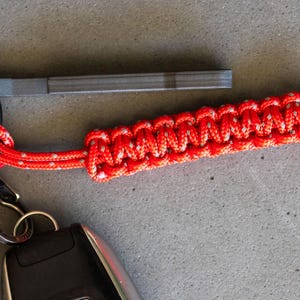 May include: A red and white paracord keychain with a gray metal fire starter attached. The keychain is attached to a set of keys.