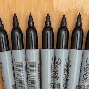 May include: A close-up of eight black Sharpie permanent markers lined up on a wooden surface. The markers have black caps and gray barrels with the Sharpie logo and barcode printed on them.