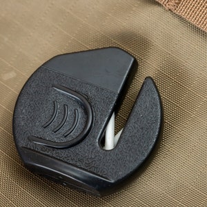 May include: A black, textured, plastic tool with a curved blade and a white, pointed insert. The tool has a rounded shape with a textured surface and a design on the front. The background is a tan, textured fabric.