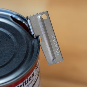 May include: A silver can opener with the text "U.S. SHELBY CO." on the side, partially open a can with a black lid. The can has the text "BEST IF USED BY AUG 28, 2025" printed on the lid.
