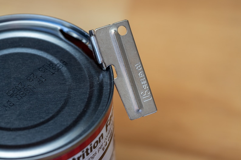 May include: A silver can opener with the text "U.S. SHELBY CO." on the side, partially open a can with a black lid. The can has the text "BEST IF USED BY AUG 28, 2025" printed on the lid.
