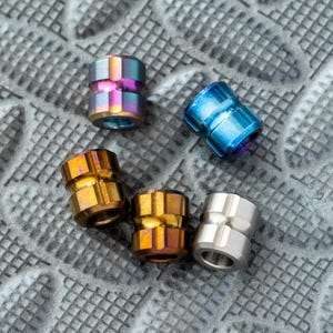 May include: A collection of five small, cylindrical beads in various metallic finishes. The beads are in shades of blue, gold, and silver, with one featuring a rainbow iridescent effect. They are arranged on a textured, gray surface.