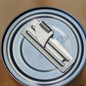 May include: A silver can opener is placed on top of a silver can. The can has a label with the text "11531717" and "AS" printed on it.