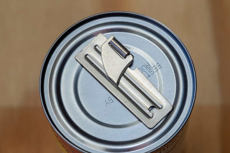 May include: A silver can opener is placed on top of a silver can. The can has a label with the text "11531717" and "AS" printed on it.