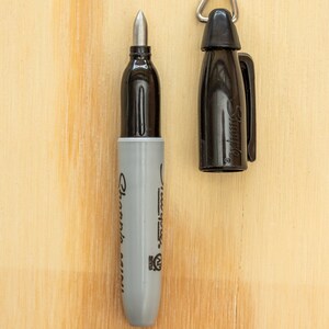 K&ouml;nnte beinhalten: Ein schwarzer Sharpie Mini Marker mit abgenommener Kappe, der die silberne Spitze zeigt. Der graue Schaft ist mit "Sharpie Mini" beschriftet. Die Kappe, mit einer silbernen Schlaufe, liegt neben dem Marker auf einer Holzoberfl&auml;che.