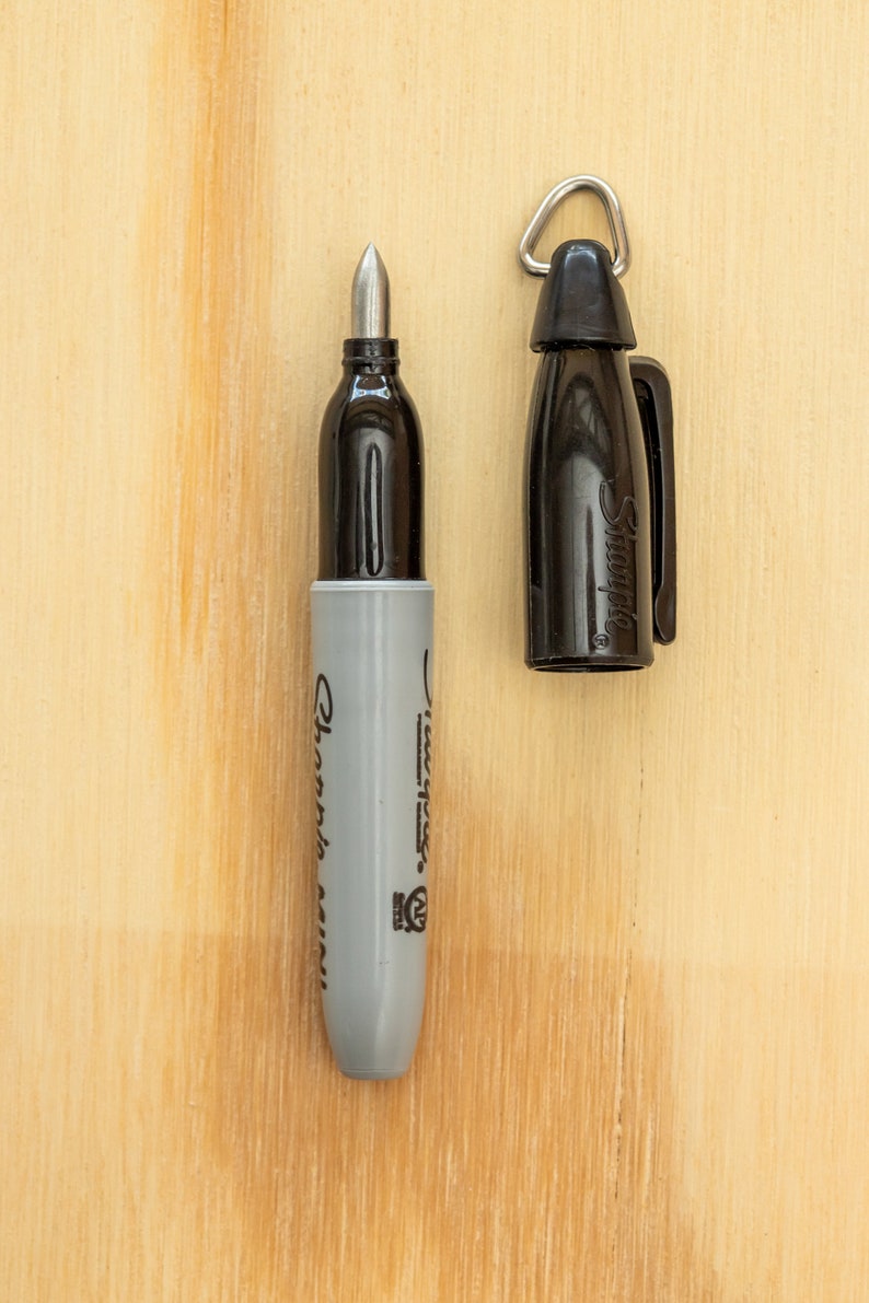 K&ouml;nnte beinhalten: Ein schwarzer Sharpie Mini Marker mit abgenommener Kappe, der die silberne Spitze zeigt. Der graue Schaft ist mit "Sharpie Mini" beschriftet. Die Kappe, mit einer silbernen Schlaufe, liegt neben dem Marker auf einer Holzoberfl&auml;che.