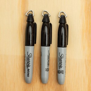 K&ouml;nnte beinhalten: Drei schwarze und graue Sharpie Mini Marker mit silbernen Schlaufen an den Kappen, vertikal angeordnet vor einem hellen Holzmaserungshintergrund. Die Marker sind mit dem Markennamen Sharpie beschriftet.