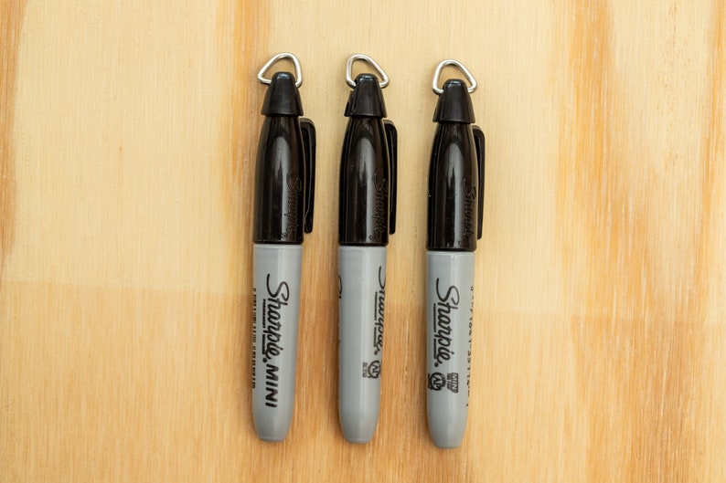K&ouml;nnte beinhalten: Drei schwarze und graue Sharpie Mini Marker mit silbernen Schlaufen an den Kappen, vertikal angeordnet vor einem hellen Holzmaserungshintergrund. Die Marker sind mit dem Markennamen Sharpie beschriftet.