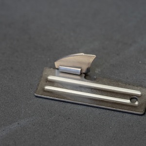 May include: A silver can opener with a small, pointed blade and a handle with a hole. The opener is lying on a gray surface.