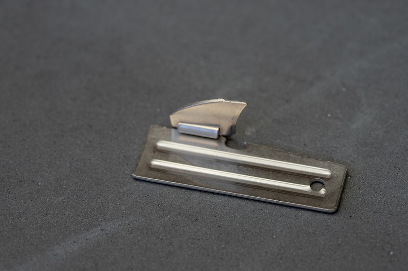 May include: A silver can opener with a small, pointed blade and a handle with a hole. The opener is lying on a gray surface.