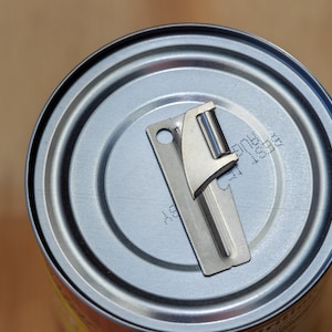 May include: A silver can opener is sitting on top of a silver can. The can has a black label with the text "BEST 12001" printed on it.