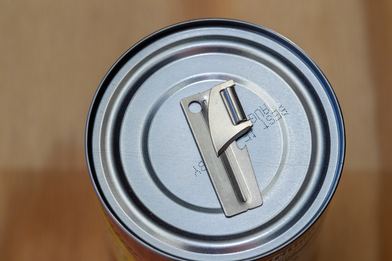 May include: A silver can opener is sitting on top of a silver can. The can has a black label with the text "BEST 12001" printed on it.