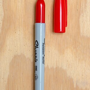 May include: A red Sharpie permanent marker with a fine point tip. The marker is on a light brown wooden surface.