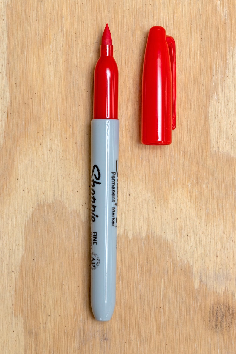 May include: A red Sharpie permanent marker with a fine point tip. The marker is on a light brown wooden surface.