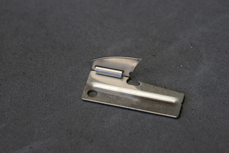 May include: A silver can opener with a small, sharp blade and a handle with a hole for opening cans.