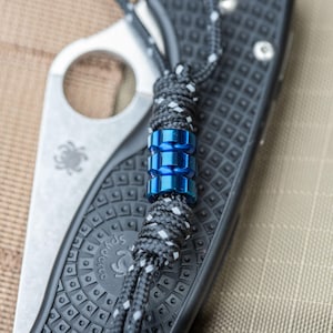 May include: A blue metal bead with a black and white cord attached to a black and silver pocket knife.