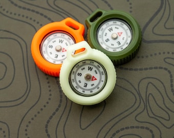 May include: Three colorful compasses: orange, green, and off-white. Each compass has a white face with black markings and a red needle. The compasses are arranged on a textured, olive-green surface with a topographic map design.