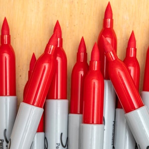May include: A close-up of a group of red permanent markers with white caps. The markers are arranged in a triangular shape, with the tips pointing upwards.