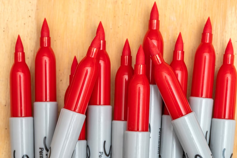 May include: A close-up of a group of red permanent markers with white caps. The markers are arranged in a triangular shape, with the tips pointing upwards.