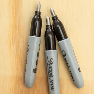 K&ouml;nnte beinhalten: Drei Sharpie Mini Permanentmarker mit grauen Geh&auml;usen und schwarzen Kappen liegen auf einer hellen Holzoberfl&auml;che. Die Marker haben silberne Spitzen und das Sharpie Mini Logo ist sichtbar.