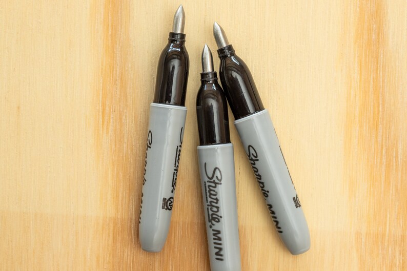 K&ouml;nnte beinhalten: Drei Sharpie Mini Permanentmarker mit grauen Geh&auml;usen und schwarzen Kappen liegen auf einer hellen Holzoberfl&auml;che. Die Marker haben silberne Spitzen und das Sharpie Mini Logo ist sichtbar.