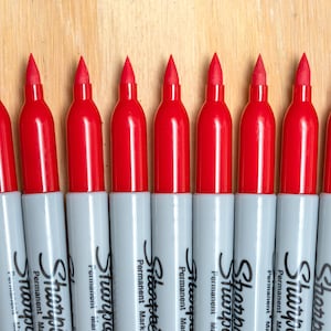 May include: A row of ten red Sharpie permanent markers lined up on a wooden surface.
